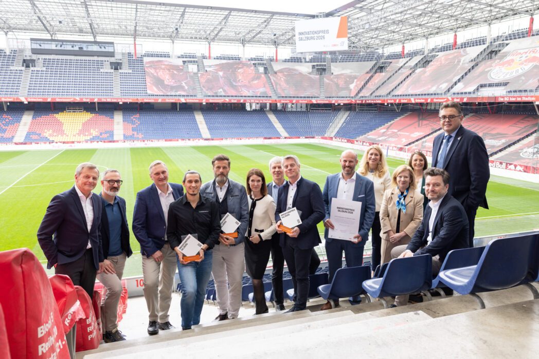 Gruppenfoto mit mehreren Personen auf der Tribühne im Stadion. Im Hintergrund auf einer großen Leinwand steht "Innovationspreis Salzburg 2026". Drei Personen in der Mitte halten einen Award, eine Person eine Urkunde.